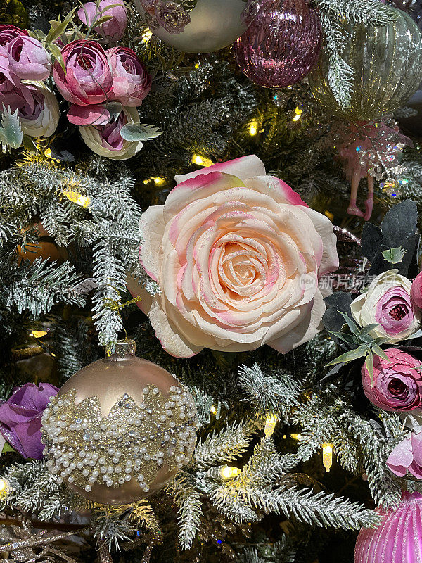 个人形象的人造，玫瑰花头饰品挂在人造圣诞树的树枝上，云杉针覆盖着人造喷雾雪，模糊的白色精灵灯的背景