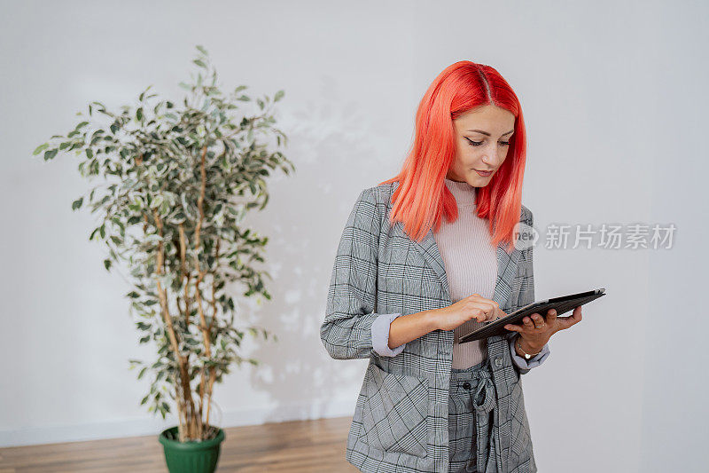 穿着优雅的女人站在公寓的空房间中间，粉红色头发的女孩房地产经纪人设计师业主手中握着平板电脑与家居家具的设计灵感