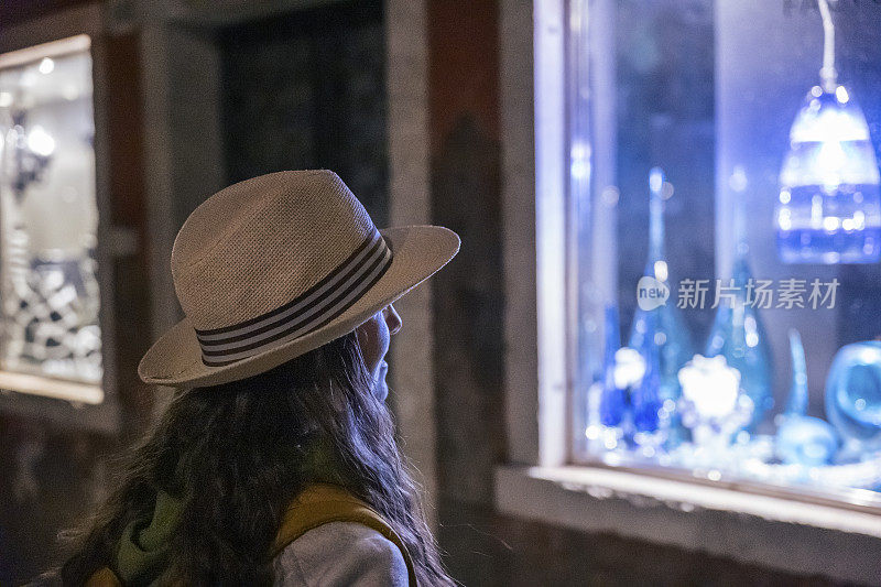 在慕拉诺岛度假时，一名女子在晚上看着商店的玻璃艺术橱窗。