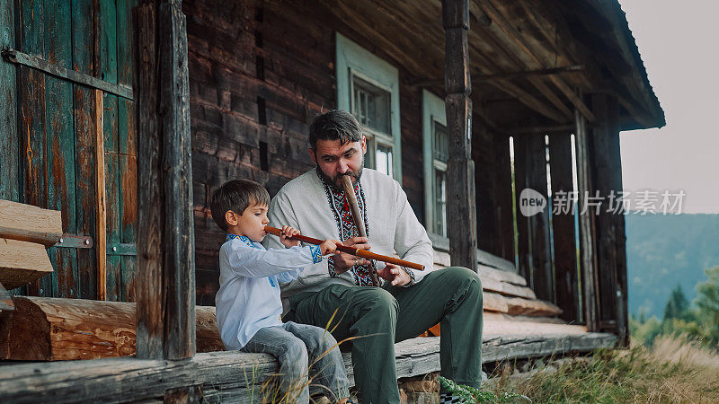小男孩和爸爸在玩木管木笛——乌克兰索皮卡。家庭二重唱，民乐概念。身穿传统刺绣衬衫的父子。他们坐在老木屋的门廊上