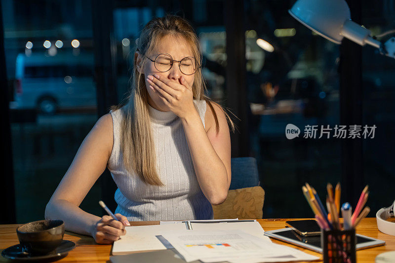 年轻的女商人在办公室的桌子上，坐在笔记本电脑和平板电脑前，感到疲惫和困倦