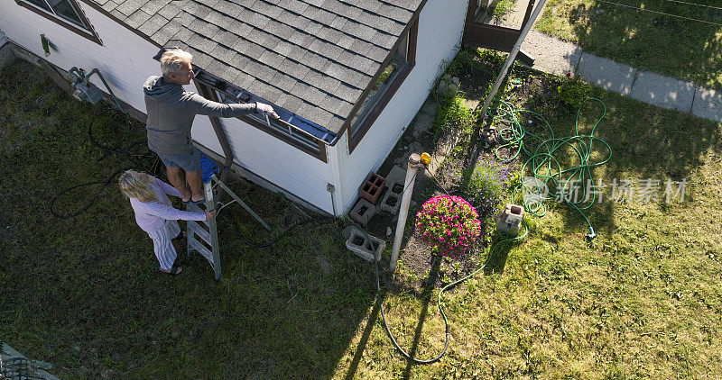 老人清理屋顶排水沟，他的同伴帮他扶梯子