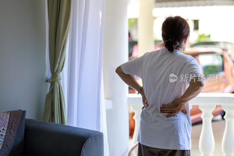 一位亚洲资深女性的后视图，疼痛地抓着她的下背部，表明她可能正在经历不适或背部疼痛。