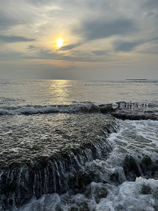 海边橙色的太阳映照在海面上，泡沫的海浪冲破绿藻和海藻覆盖的岩石，岩石潮汐池，潮汐反射，太阳落在涟漪的水面上，复制空间