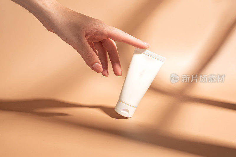 女人的手摸着一筒白色奶油的模型。未标示的化妆品包装在阳光下的米色背景。护肤概念。强烈的阴影