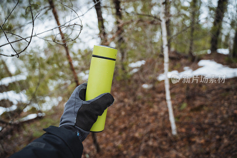 一个绿色的保温瓶以春天的森林为背景，一个男人戴着手套拿着一个保温瓶。