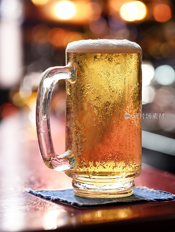 啤酒在玻璃大酒杯的特写图像覆盖了凝结与起泡的气泡形成泡沫的顶部啤酒，杯杯杯杯杯杯在木质吧台，酒吧设置背景，重点在前景
