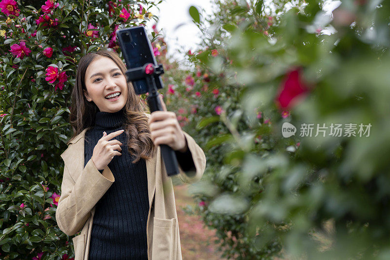 韩国一名微笑的旅游博主网红在韩国济州岛的玫瑰花园公园拍摄冬季视频，她正在用智能手机为她的粉丝直播