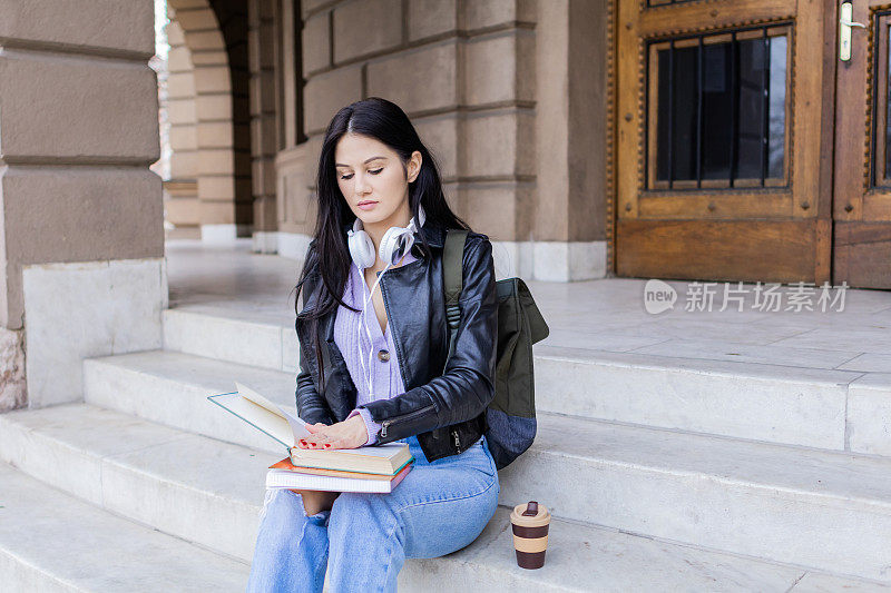 年轻漂亮的女大学生背着双肩包坐在大学楼梯上。她戴着耳机，穿着皮夹克，等待着大学课程的开始。