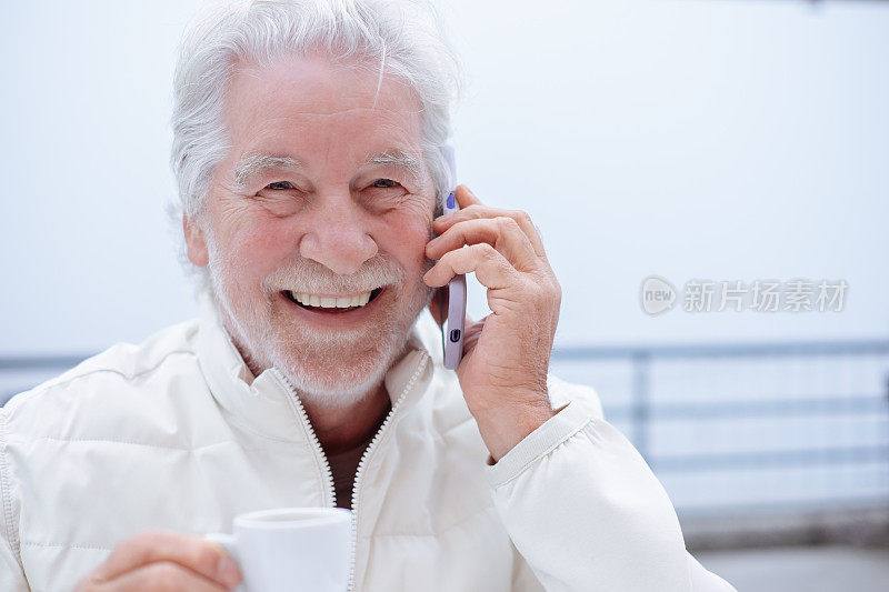 开朗迷人的白发老人坐在户外的咖啡桌旁，用智能手机聊天，喝着浓咖啡杯休息一下