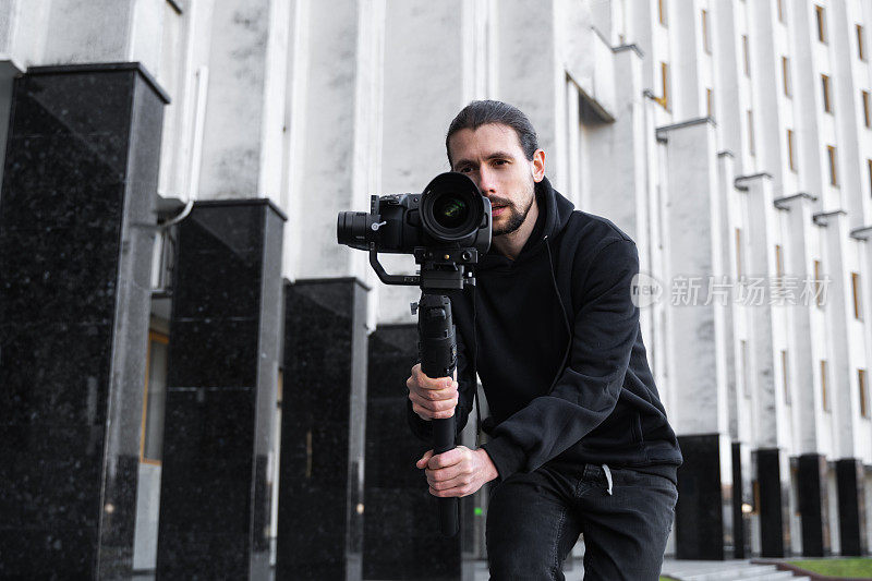 年轻的专业摄像师手持专业摄像机，安装在三轴框架稳定器上。专业设备有助于制作高质量的视频而不摇晃。一个穿着黑色连帽衫的摄影师正在录像。