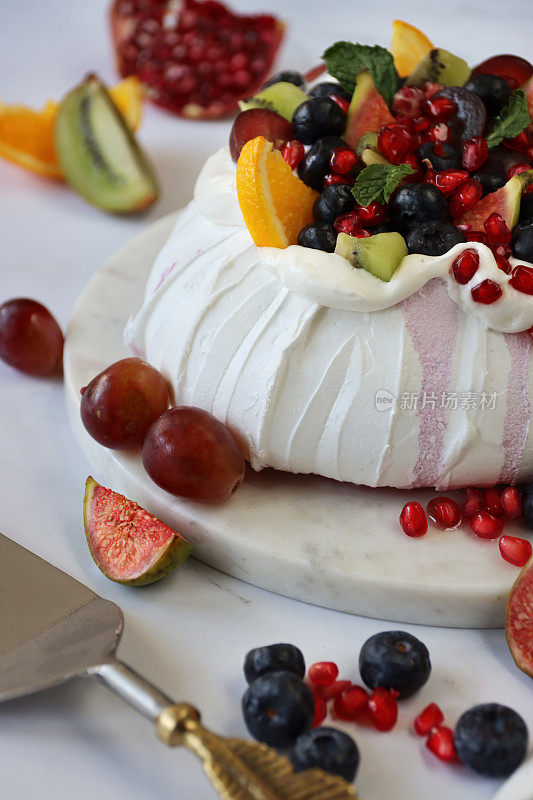 这是一个用新鲜水果(包括橙子、石榴籽、蓝莓、葡萄、猕猴桃和无花果)填满的酥皮的特写，放在一个大理石蛋糕架上，旁边是茶巾，金属蛋糕服务员，以大理石效果为背景，聚焦前景