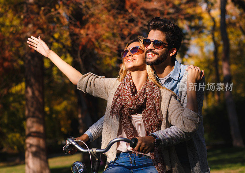快乐的年轻夫妇在阳光明媚的秋天在公园里骑自行车。