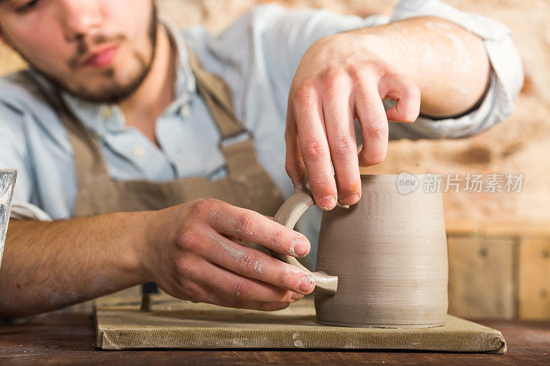 陶艺、石器、陶艺的艺术概念——工匠的双手将生土制成的杯子和把手连接起来，大师的手指摆弄着一块块陶土，男性坐在桌子后面的工作间里