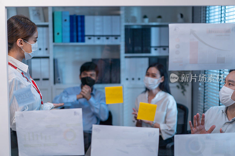 一群亚洲商人和妇女戴着口罩，在办公室开会。员工群策群力，以团队的形式工作，在纸上逐点计划和讨论项目。现代同事或同事的公司。