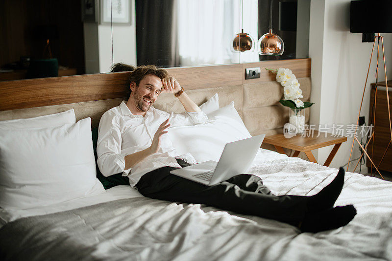 商人躺在酒店的床上，用笔记本电脑和家里的家人进行视频通话