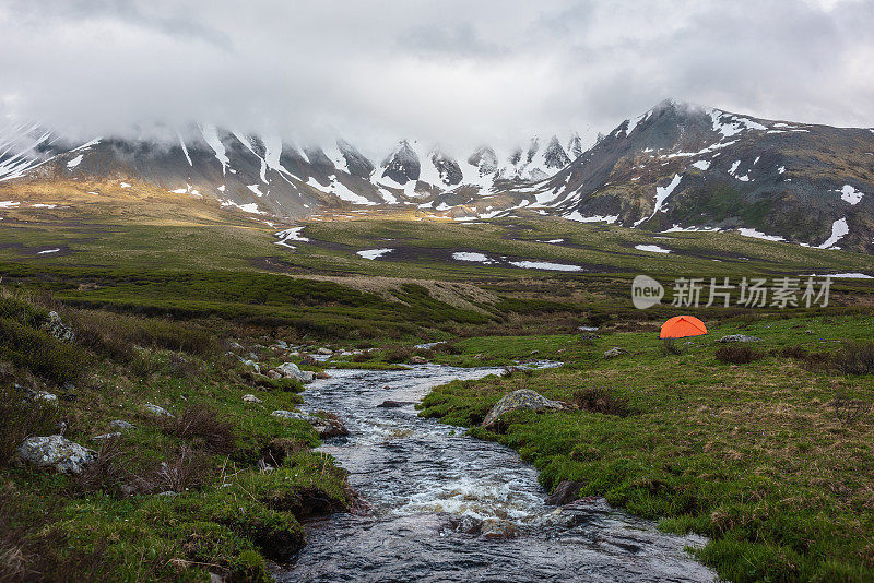 橙色帐篷靠近清澈的小溪，在低低的雨云中可以看到山脉。戏剧性的高山景观与山溪和雪山在灰色的低云。冻土带阴森的景色。