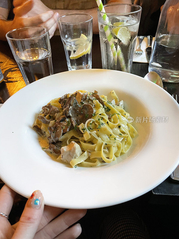 图片的餐厅场景的餐桌设置的奶油鸡肉和蘑菇意大利面条食谱在意大利餐厅，吃出意大利面菜在白色的盘子，喝的杯子，不认识的人在背景