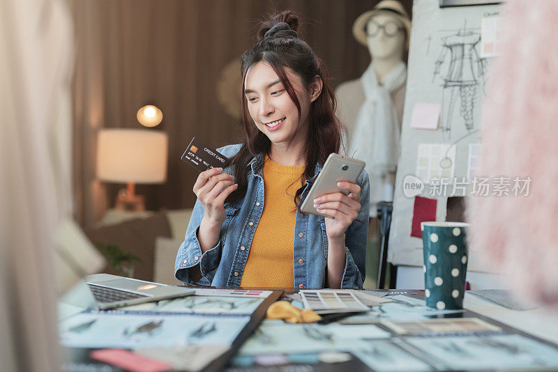 亚洲女性成年女性创意时尚服装设计师微笑，手拿智能手机和信用卡在线购买和购物在她的工作室办公室