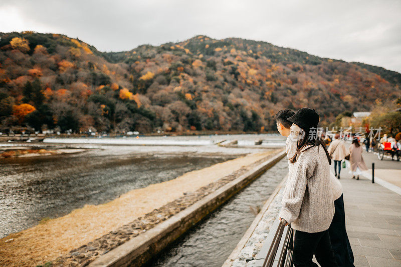 一对年轻的亚洲游客夫妇正在欣赏以河和山为背景的美丽的秋景。