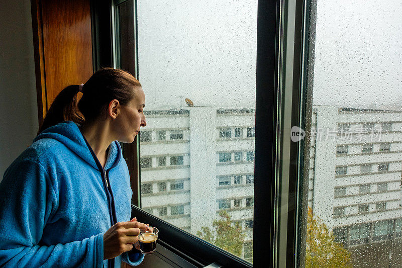 下雨天，一个手里拿着咖啡的女人望着窗外