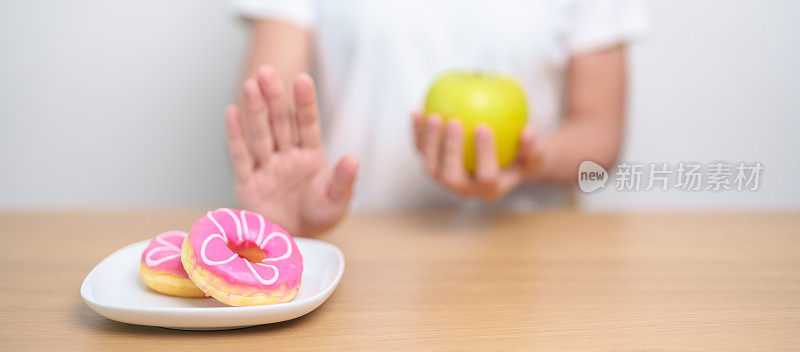 女性手持青苹果而拒绝甜甜圈，女性健身在健康水果和不健康的垃圾食品之间选择。节食控制，体重减轻，肥胖，饮食方式和营养