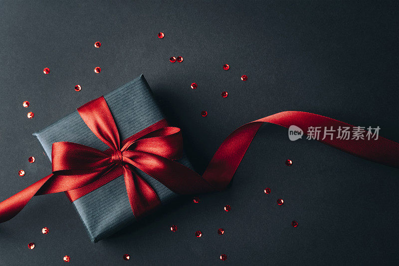 礼品盒与红色蝴蝶结黑色背景与红色五彩纸屑。圣诞节、生日、节日的概念。俯视图，平铺