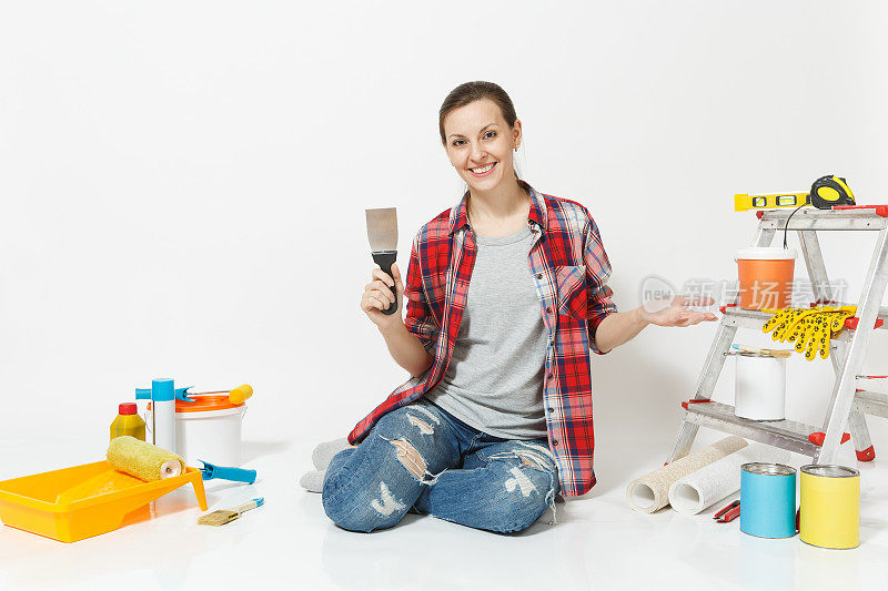 穿着便服的女子坐在地板上用油灰刀，仪器为装修公寓房间隔离在白色的背景上。壁纸配件用于粘漆工具。修理家里的概念。