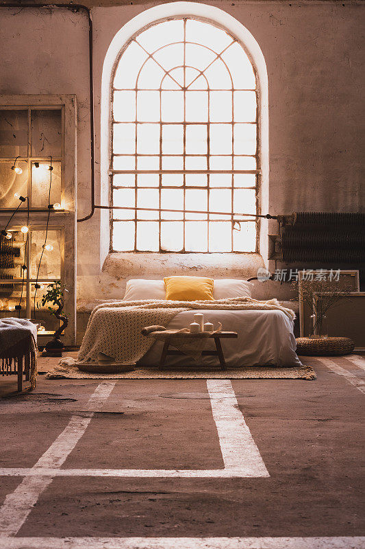 宽敞的工业卧室室内，床上方的窗户配有灯和带毯子的木椅。真实的照片
