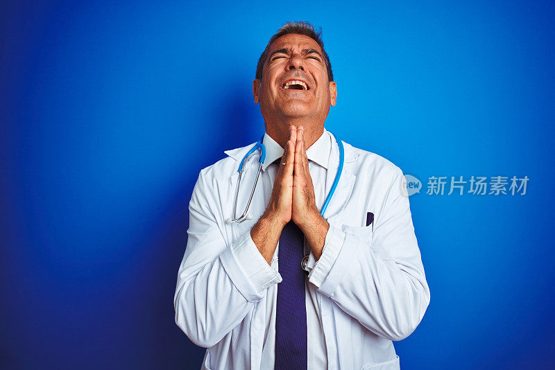 英俊的中年医生男子戴着听诊器在孤立的蓝色背景乞求和祈祷与希望的表情在脸上非常激动和担心。请求宽恕。宗教的概念。