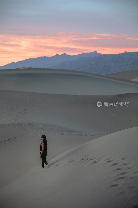 美丽的女人站在沙丘上与脚印在相机回望生机勃勃的日落-美丽的橙色天空-冒险背景