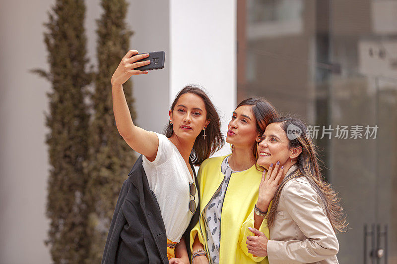 早晨阳光下，三位拉丁美洲女同事站在办公大楼外:社交媒体的自拍时间到了;他们在网络媒体前摆姿势——显然这是他们千禧一代正常生活的一部分。