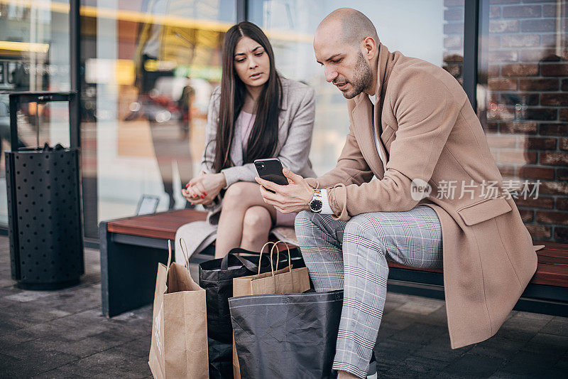 一男一女拿着购物袋坐在长椅上玩手机