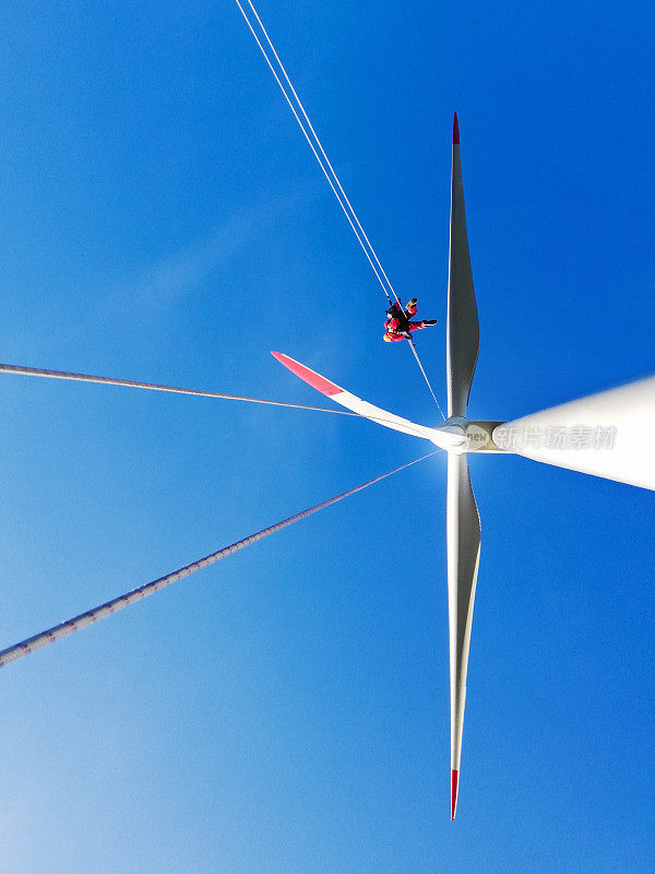 从底部看风力发电机和工业攀登者-绳索访问技术人员从绳索上的叶片下降，晴朗的天空和阳光明媚的一天。
