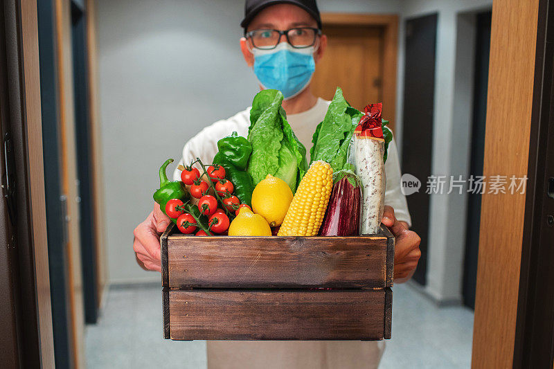 带防护口罩的送餐员手持纸袋，携带食物进入入口。快递员把装有新鲜蔬菜和水果的盒子交给顾客