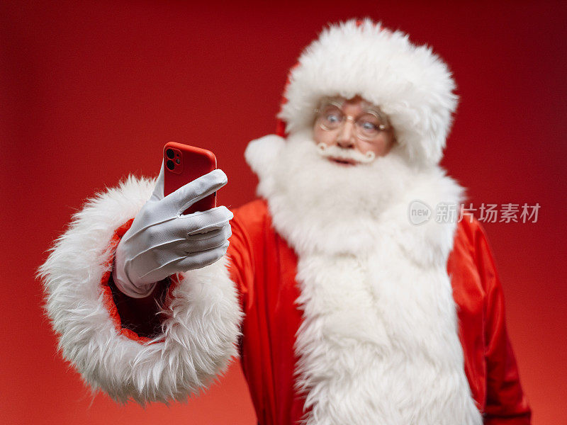 圣诞老人惊讶地看着他的手机，站在鲜红色的背景上，