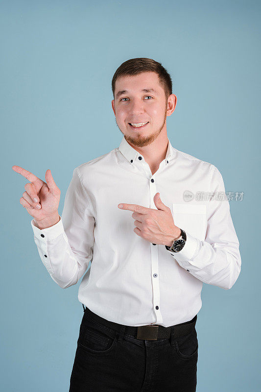 一个25-29岁的男人，穿着白衬衫，留着胡子，双手站在一张蓝色背景的垂直照片上，做着食指的手势