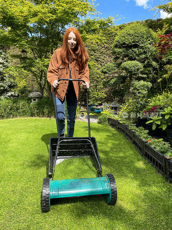 有吸引力的特写图像，红头发的年轻女子做园艺家务，园丁修剪草坪与推割草机，圆筒割草机，阳光明媚的日子，重点在前景