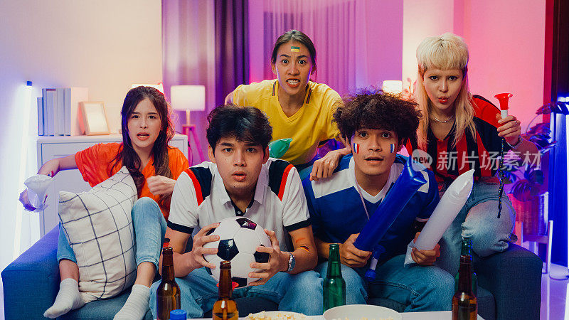 晚上，一群快乐的亚洲青少年坐在沙发上看电视上的足球比赛。