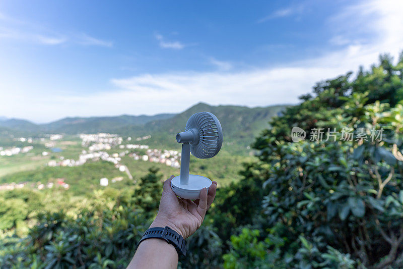 一只手拿着小电风扇，背景是乡村的蓝天白云