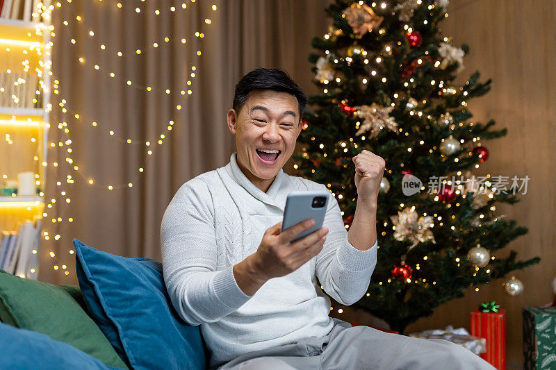 一名亚洲男子坐在家里一棵9岁的圣诞树旁玩手机