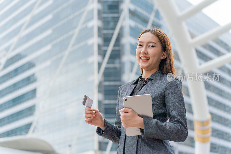 在一个商业区，一名身穿灰色西装的年轻亚洲女商人在使用她的信用卡通过她的数字平板电脑进行在线交易时自信地微笑着