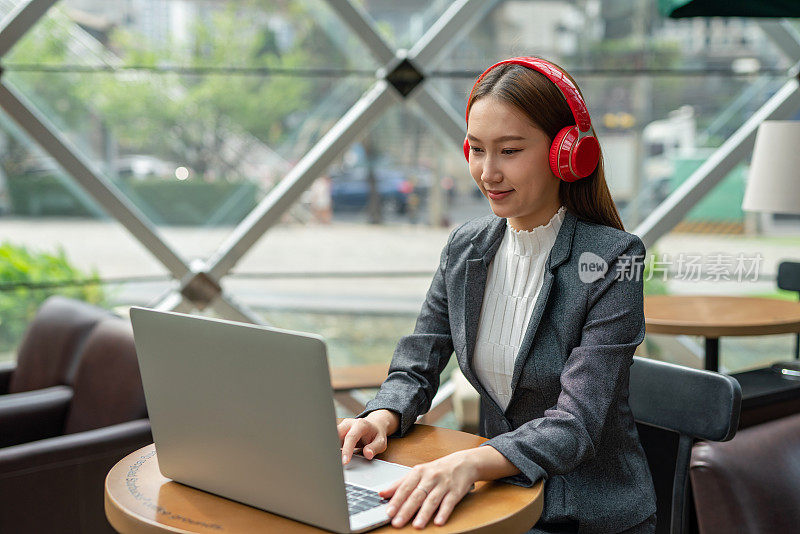 混合式工作风格。年轻的亚洲女性成年人与笔记本电脑在咖啡馆工作。戴着红色耳机的女商人