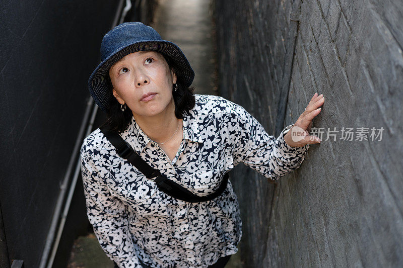 一名戴着帽子的日本妇女站在两栋建筑之间的狭窄小路上，抬起头来