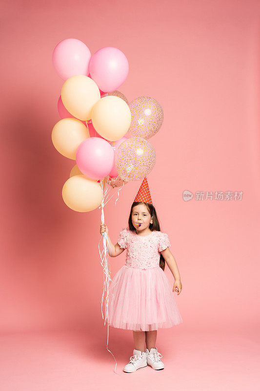 快乐的庆祝生日派对与迷人的可爱的小女孩在薄纱衣服微笑着对着相机孤立在粉红色的背景气球飞行庆祝。迷人的微笑，表达幸福