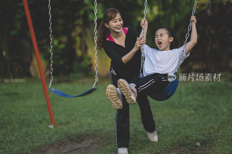 一位亚洲华人母亲在周末的闲暇时间推着女儿在公园里玩秋千
