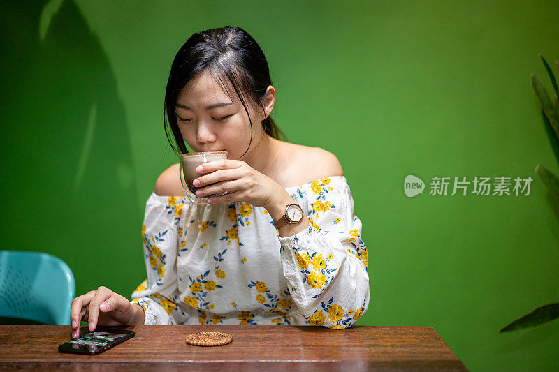 年轻的亚洲女子一边喝咖啡一边用智能手机