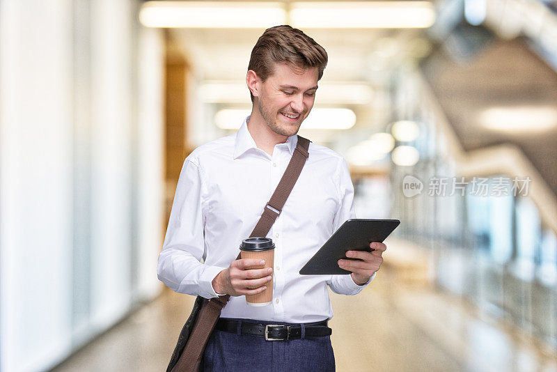 男性企业家站在办公室散焦背景前，肩包和拿包，使用触摸屏