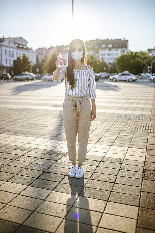 一名年轻女子戴着外科口罩，手上涂着洗手液，在封锁期间站在空荡荡的城镇广场上