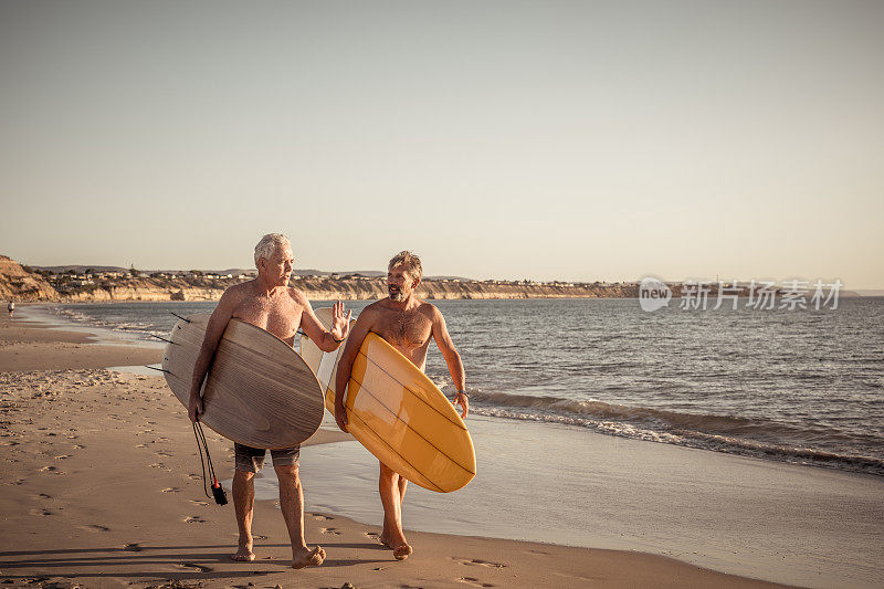 两个成熟的男人带着冲浪板在美丽的海滩上散步，享受着天堂和退休生活。有吸引力的老年人朋友有乐趣的冲浪。在真正积极健康的人身上。